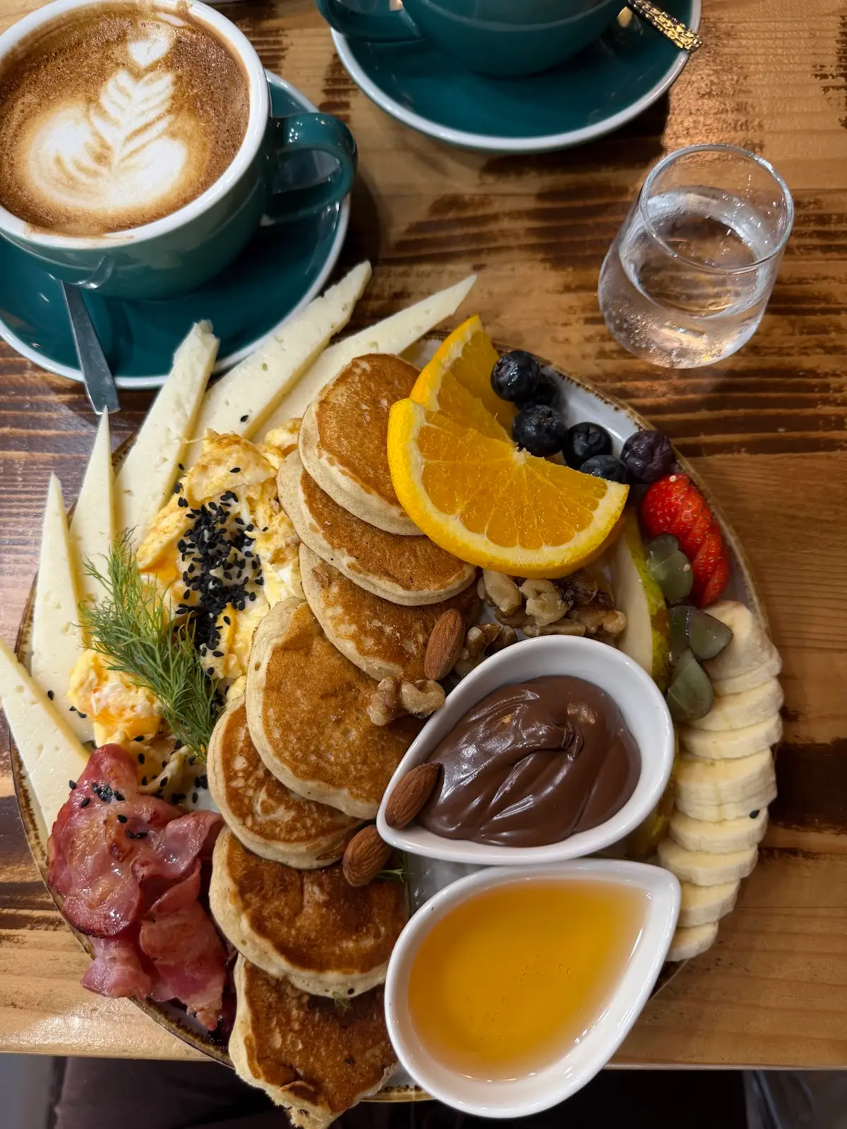 Tortitas con fruta, miel y chocolate, un cortado en taza verde azulada, vistos desde arriba