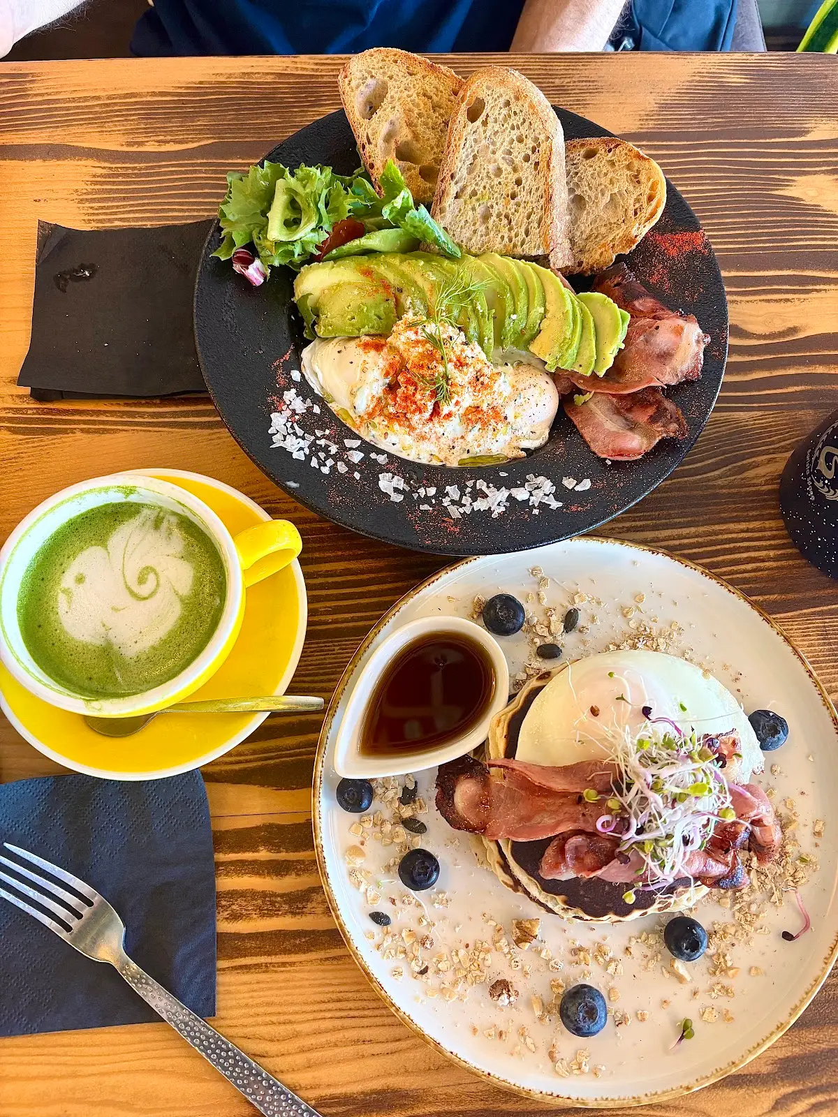 Tosta de aguacate en plato de piedra negra, matcha latte en taza amarilla y tortitas con arándanos