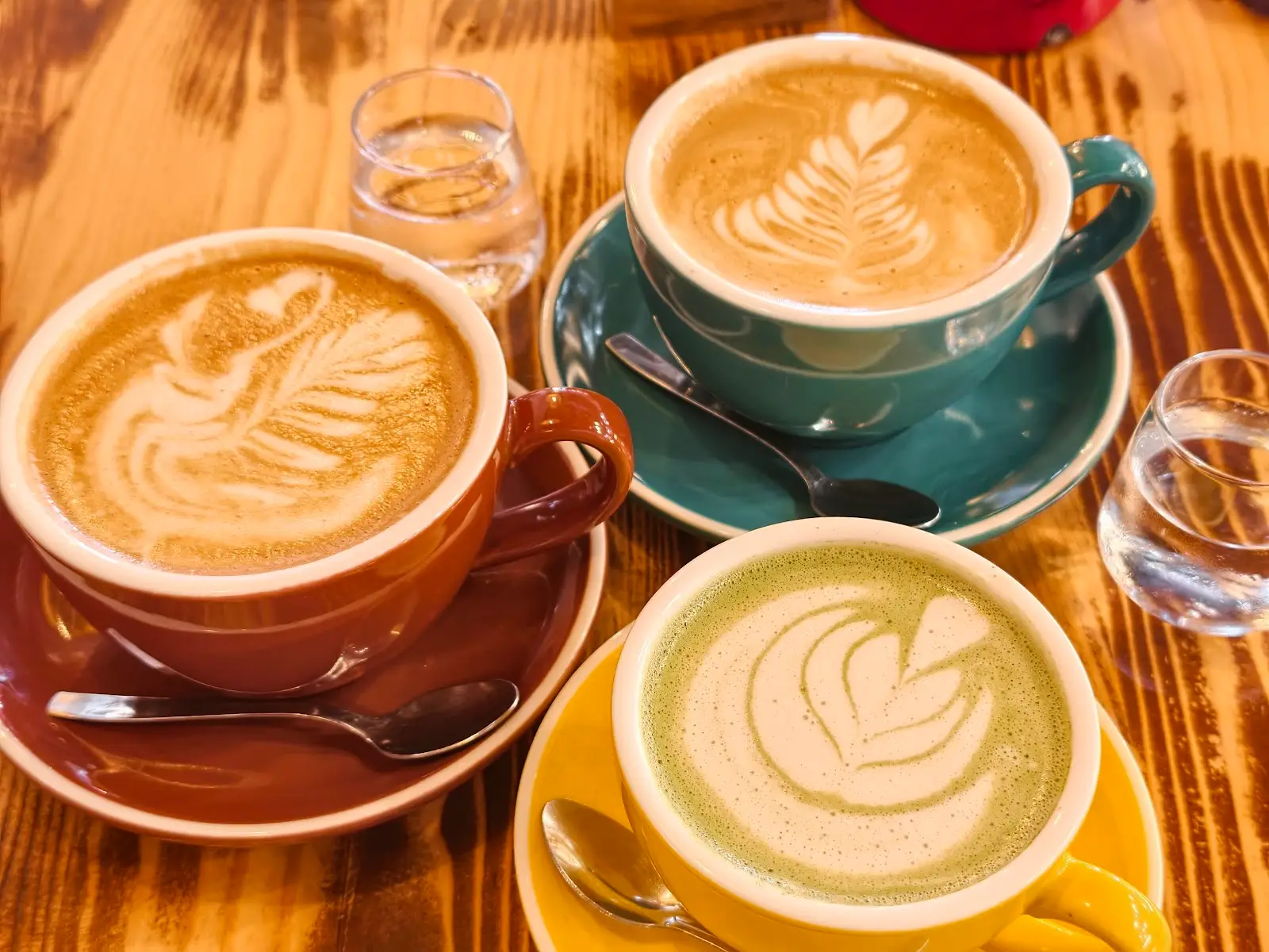 Tres cafés con dibujo en la espuma en tazas de color teja, verde azulado y mostaza
